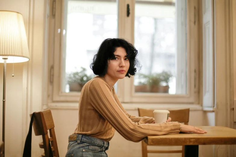 a woman sitting at a table with a cup of coffee, inspired by Elsa Bleda, pexels contest winner, realism, finn wolfhard, beautiful iranian woman, brown shirt, low quality photo