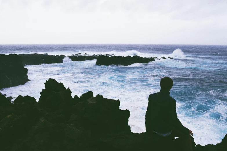 a man sitting on top of a rock next to the ocean, an album cover, pexels contest winner, minimalism, azores, ((waves, silhouette of man, looking sad