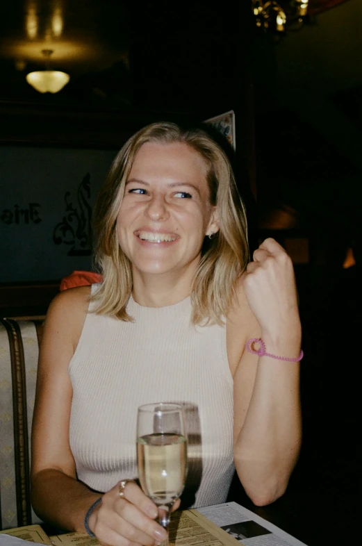 a woman sitting at a table with a glass of wine, smiling for the camera, taken in the late 2000s, slightly pixelated, a blond