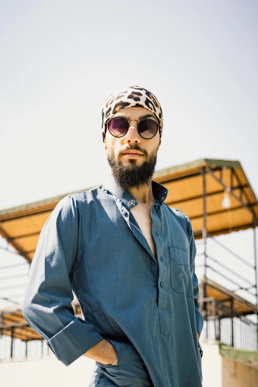 a man with a beard standing in front of a building, an album cover, by Giorgio De Vincenzi, unsplash, wearing a turban, blue sky, jewish young man with glasses, color vintage