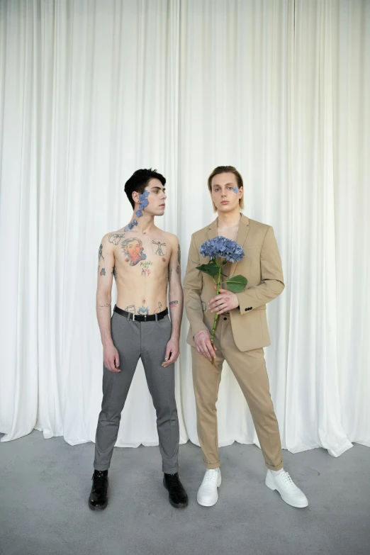 a couple of men standing next to each other, an album cover, by Ellen Gallagher, aestheticism, full-body tattoos, there are flowers, nonbinary model, tan suit