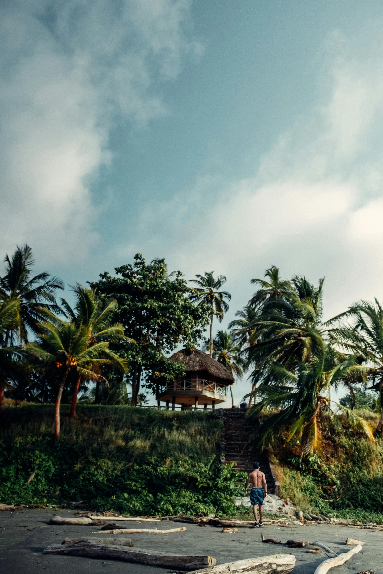 a man riding a surfboard on top of a sandy beach, tribe huts in the jungle, house on a hill, banana trees, cinematic photograph