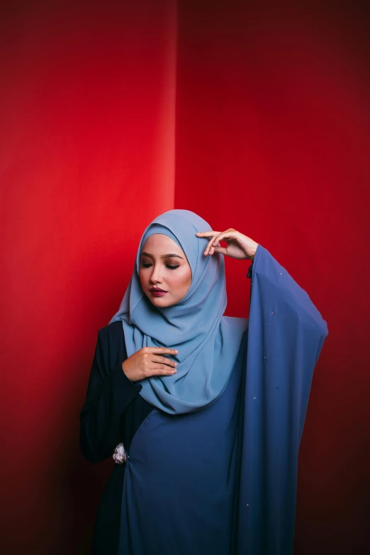 a woman in a blue hijab standing against a red wall, pexels contest winner, hurufiyya, grey clothes, smooth in _ the background, pose 4 of 1 6, shawl