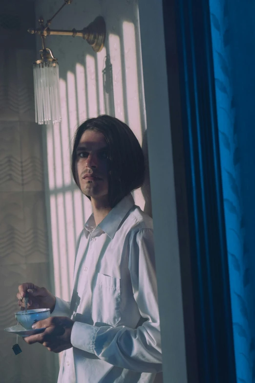 a man standing in front of a window holding a cup, an album cover, inspired by Elsa Bleda, unsplash, surrealism, avan jogia angel, blue room, adam driver, ( ( theatrical ) )
