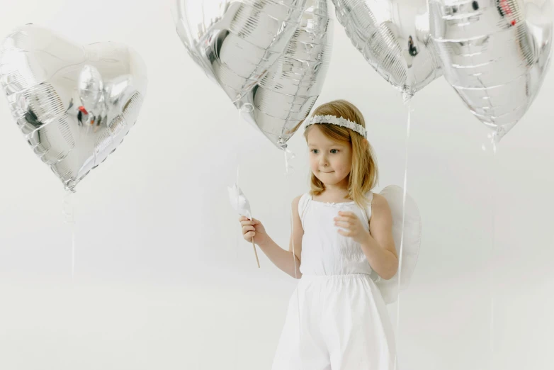 a little girl standing in front of a bunch of balloons, by Marie Angel, pexels contest winner, white and silver, girl with angel wings, on a white table, silver accessories