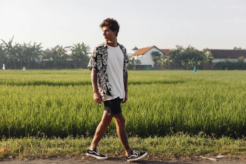 a man walking across a dirt road next to a lush green field, a portrait, trending on pexels, happening, bra and shorts streetwear, batik, white and black clothing, avatar image