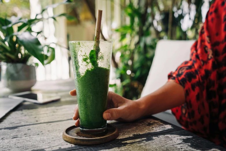 a person sitting at a table with a glass of green smoothie, by Emma Andijewska, trending on unsplash, warm weather, 🦩🪐🐞👩🏻🦳, an abandoned, moringa juice