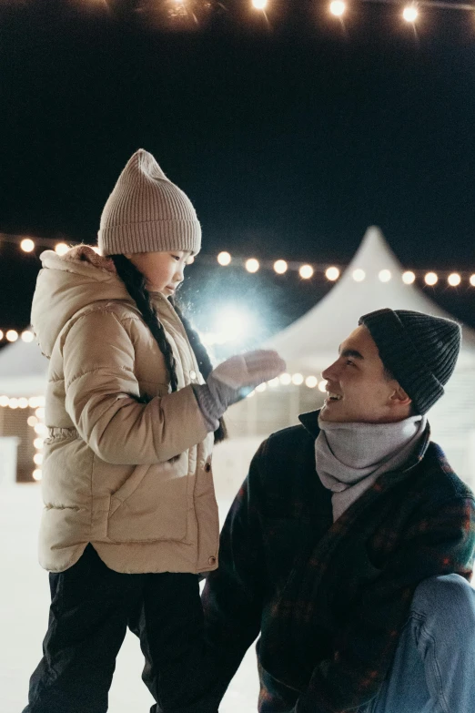 a man standing next to a woman on top of a snow covered ground, fairy lights, father with child, beanie, promo