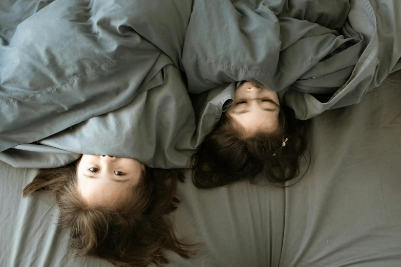 a couple of women laying on top of a bed, pexels contest winner, surrealism, two identical symmetrical eyes, cloak covering face, children, gray