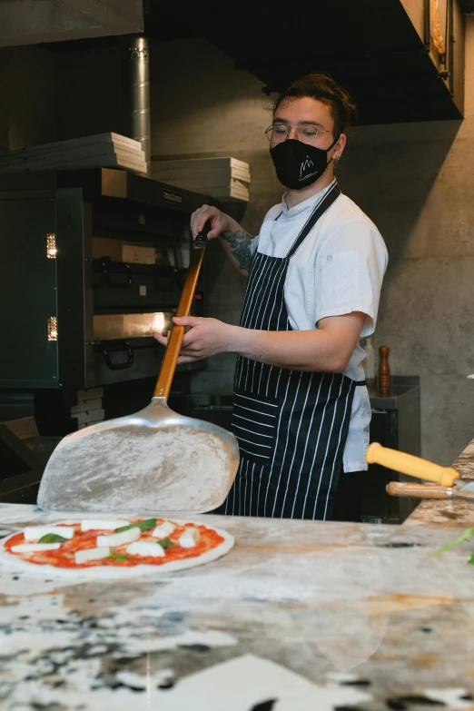 a man that is standing in front of a pizza, holding a knife, wearing facemask, working hard, spatula