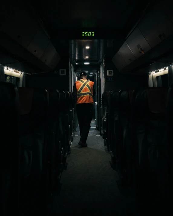 a man in an orange vest walking down the aisle of a train, by Alison Geissler, pexels contest winner, out in the dark, public bus, maintenance, hivis