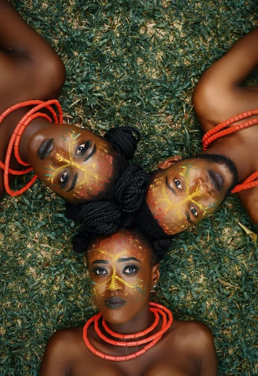 a group of women laying on top of a lush green field, an album cover, by Chinwe Chukwuogo-Roy, pexels contest winner, afrofuturism, tribal facepaint, detailed symmetrical faces, orange pupils, photograph from above