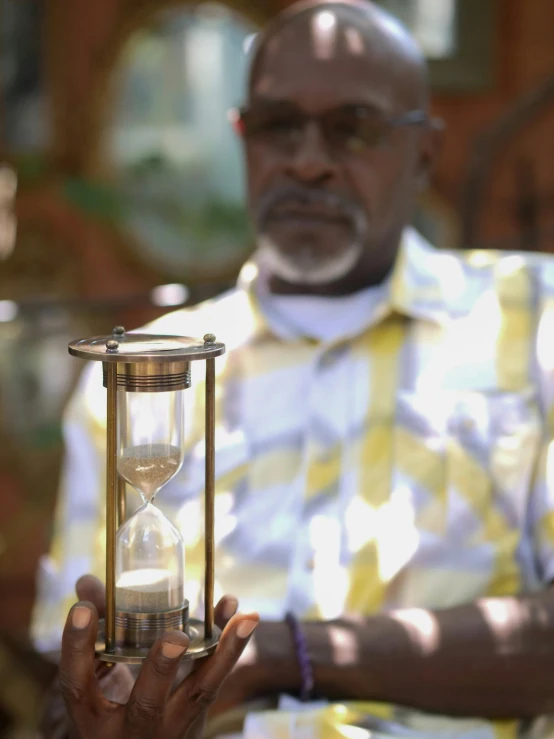 a man sitting in a chair holding a hourglass, by Robert Bain, pexels contest winner, hurufiyya, bahamas, video still, community celebration, medium close up shot