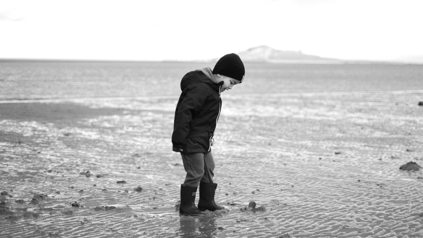 a little boy standing on top of a sandy beach, a black and white photo, inspired by Vija Celmins, pexels, walking on ice, skye meaker, standing in a shallow river, pondering