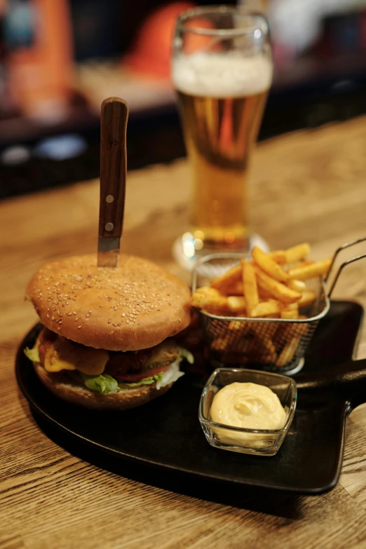 a burger sitting on top of a black tray next to a glass of beer, by Adriaen Hanneman, daily specials, thumbnail, lounge, b - roll