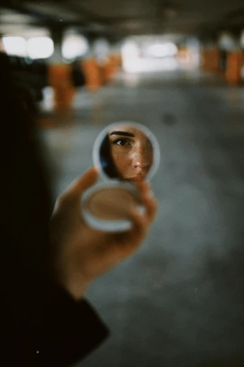 a person holding a mirror in a parking garage, a picture, inspired by Elsa Bleda, pexels contest winner, hyperrealism, small round face, with anamorphic lenses, soft oval face, small in size
