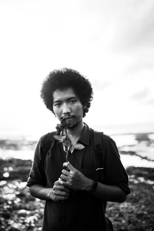 a man standing on a beach holding a flower, a black and white photo, by Basuki Abdullah, long afro hair, ((portrait)), single light, song nan li