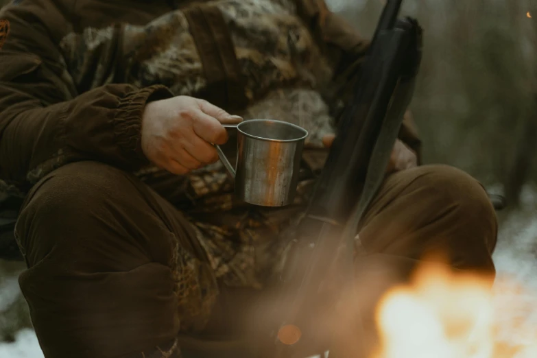 a man sitting next to a fire holding a cup, hunters gear, thumbnail, detail shot, aesthetic shot