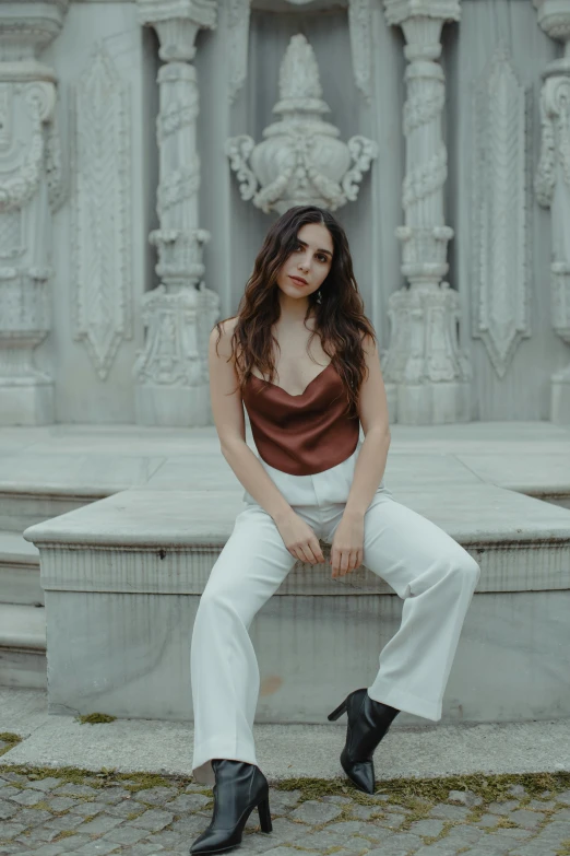 a woman sitting on a bench in front of a fountain, an album cover, inspired by Elsa Bleda, trending on pexels, baroque, white pants, ana de armas portrait, maroon, white marble buildings