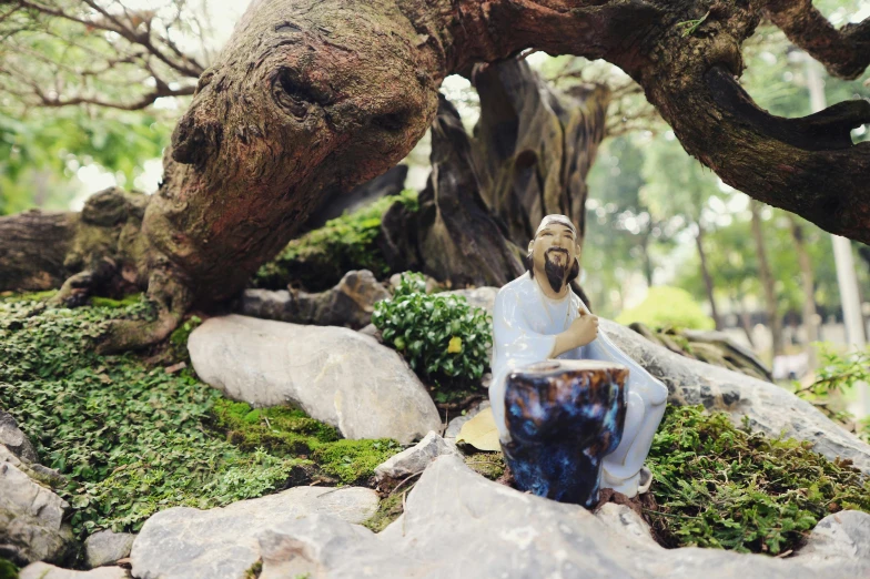 a statue of a man sitting on a rock, a statue, unsplash, environmental art, blue and pink bonsai tree, jesus of nazareth, resin and clay art, overturned chalice