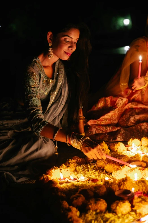 a couple of women sitting next to each other in front of a fire, hindu ornaments, glowing gold embers, blessing the soil at night, floating candles