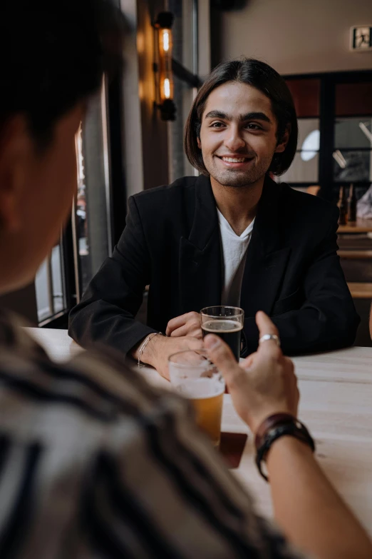 a couple of people that are sitting at a table, a man wearing a black jacket, social encounters, holding beer, smiling at each other