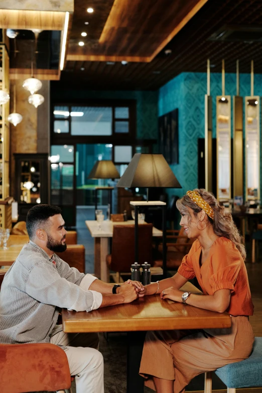 a man and woman sitting at a table in a restaurant, tanned ameera al taweel, reaching out to each other, gentleman's club lounge, millennial vibes