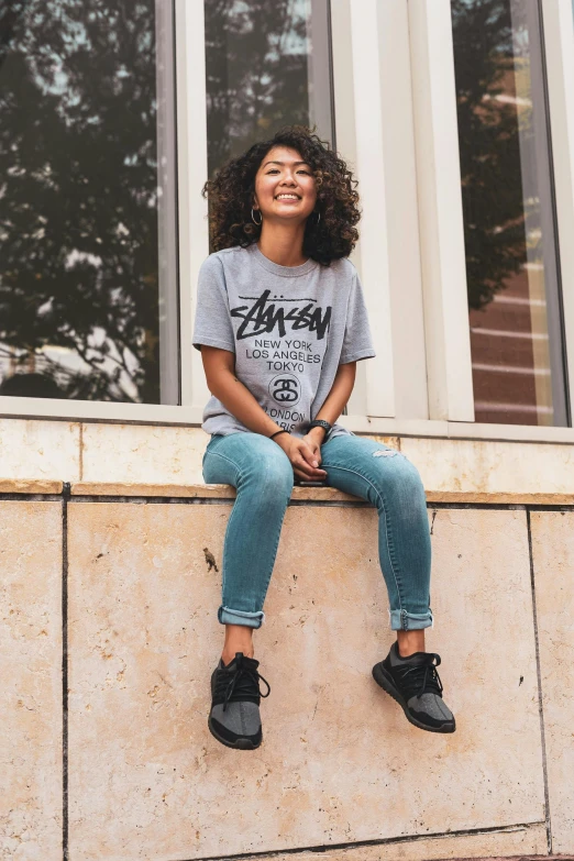 a woman sitting on a ledge in front of a window, inspired by Washington Allston, trending on unsplash, graffiti, gray shirt, young asian girl, smiling down from above, official store photo