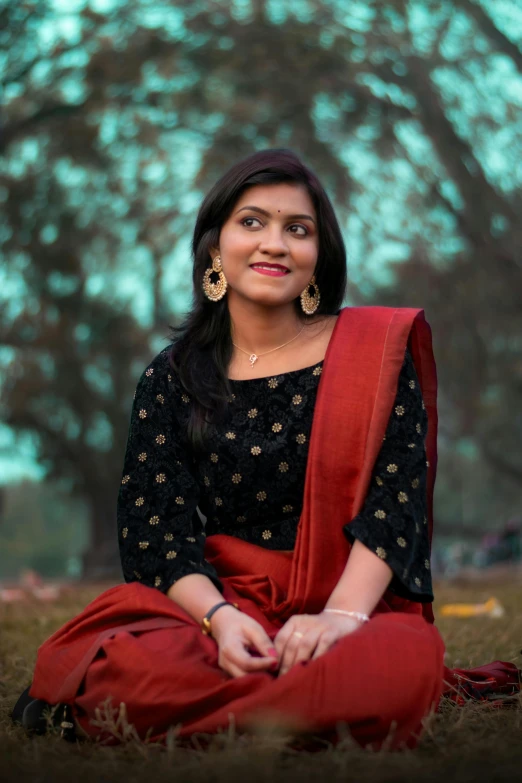 a woman sitting on the ground in a red sari, an album cover, pexels contest winner, at a park, headshot profile picture, assamese, wearing a black and red suit