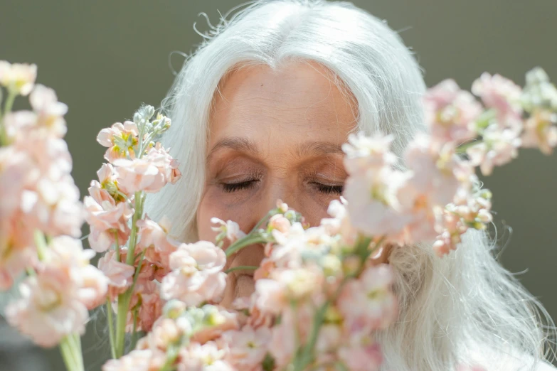 a woman smelling flowers with her eyes closed, by Ellen Gallagher, trending on unsplash, elderly greek goddess, long silver hair, in shades of peach, photoshoot for skincare brand