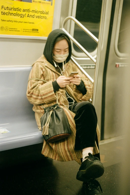 a woman sitting on a subway looking at her phone, inspired by Li Di, neo-figurative, surgical mask covering mouth, brown clothes, non-binary, wearing robe