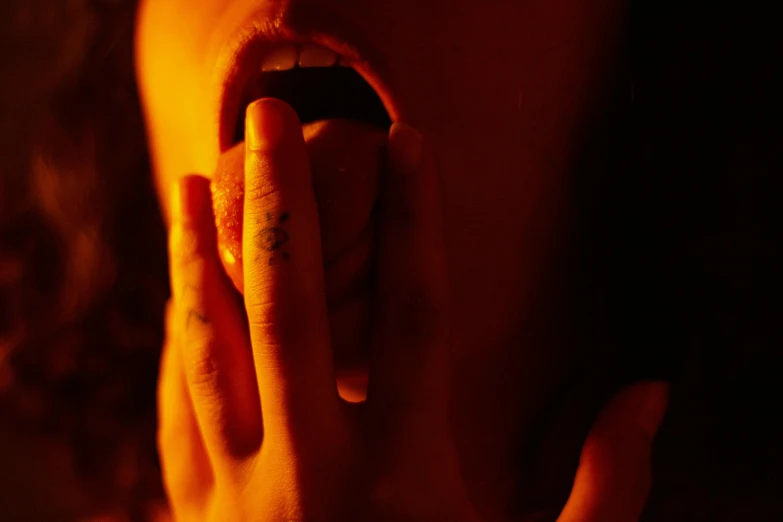 a close up of a person holding their hand to their mouth, inspired by Elsa Bleda, realism, orange and red lighting, music video, large tongue, hellish scene