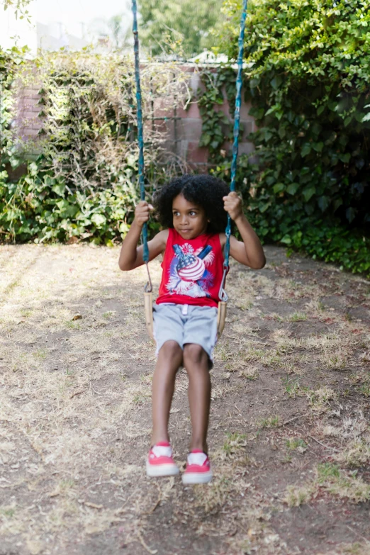 a little girl that is sitting on a swing, tan skin a tee shirt and shorts, red and blue garments, in a garden, hero shot