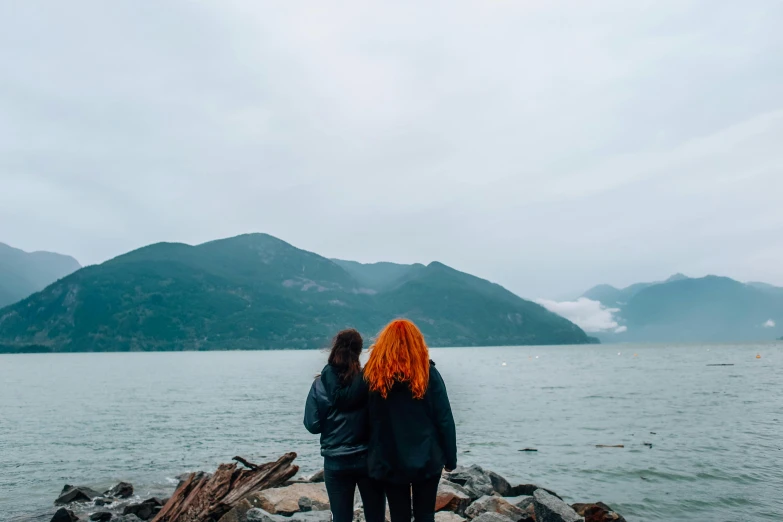 two people standing on rocks in front of a body of water, lesbian, background image, vancouver, high quality image”