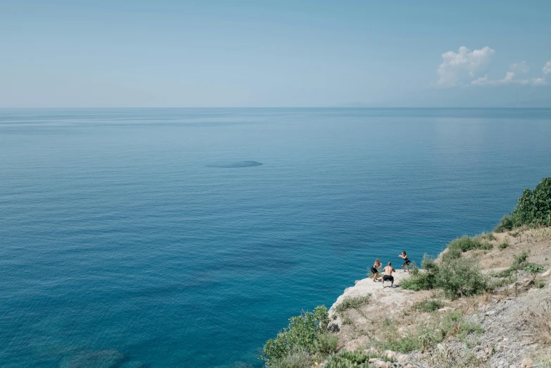 a group of people sitting on top of a cliff next to the ocean, alexandros pyromallis, blue sea, tranquillity, nature photo