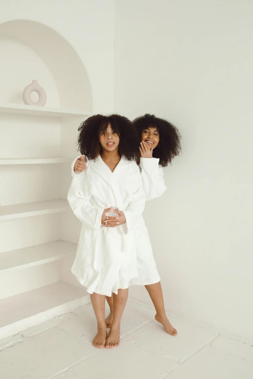 a couple of women standing next to each other, wearing a white robe, photoshoot for skincare brand, curly afro, inside white room