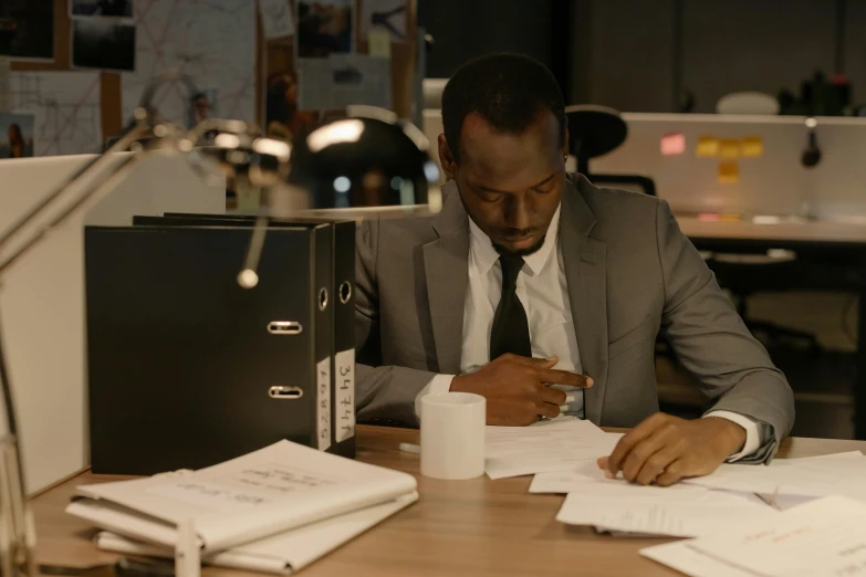 a man sitting at a desk writing on a piece of paper, pexels contest winner, private press, still image from tv series, osborne macharia, wearing dark grey suit, busy night