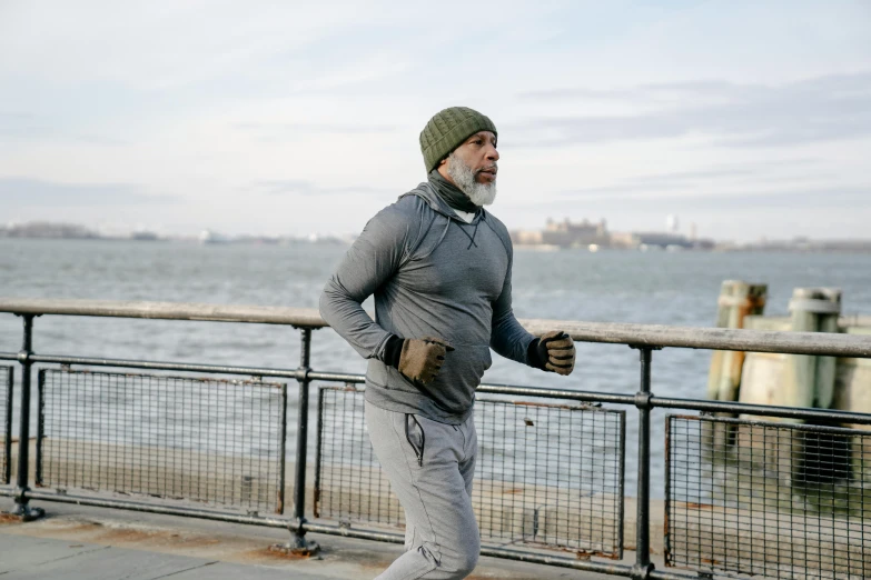 a man running on a sidewalk next to a body of water, he also wears a grey beanie, old gigachad with grey beard, compression, brooklyn