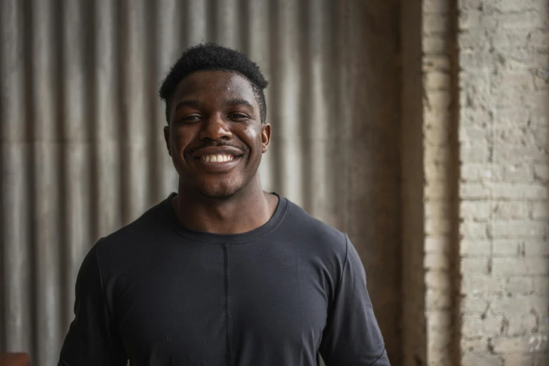 a man standing in front of a brick wall, large black smile, local gym, david uzochukwu, lachlan bailey