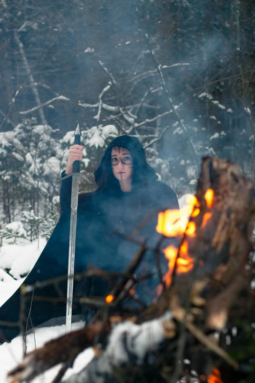 a woman standing next to a fire in the snow, wearing black medieval robes, hold sword in the forest, slide show, multiple stories