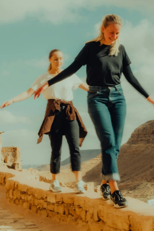 a couple of women standing on top of a stone wall, trending on pexels, happening, walking in the desert, jeans and t shirt, in style of petra collins, mid air