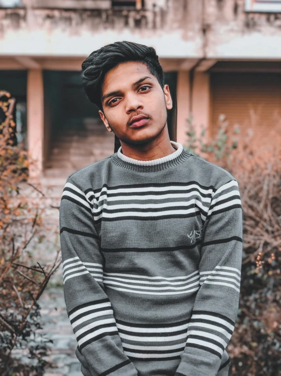 a young man standing in front of a building, an album cover, inspired by Saurabh Jethani, pexels contest winner, realism, striped sweater, headshot profile picture, dressed in a gray, ((portrait))