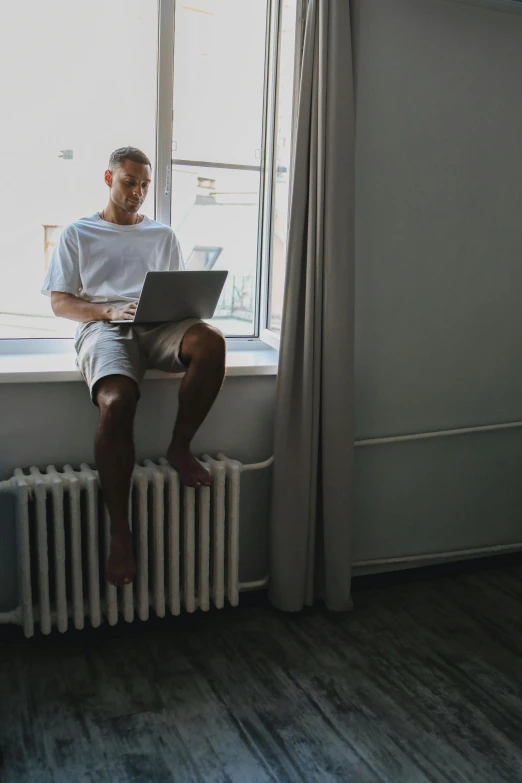 a man sitting on a radiator using a laptop, by Adam Rex, pexels contest winner, wearing shorts, window in room, alex kanevsky, luka mivsek