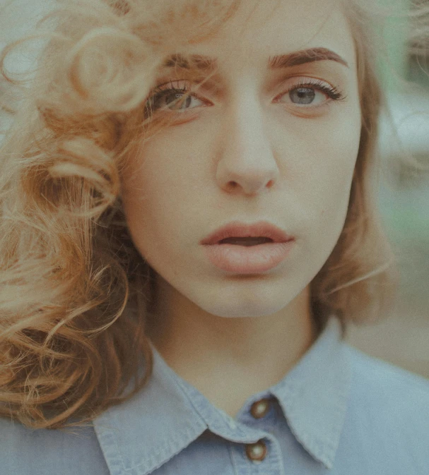 a woman with blonde hair wearing a blue shirt, inspired by Elsa Bleda, pexels contest winner, antipodeans, her face framed with curls, human staring blankly ahead, sunfaded, big nostrils