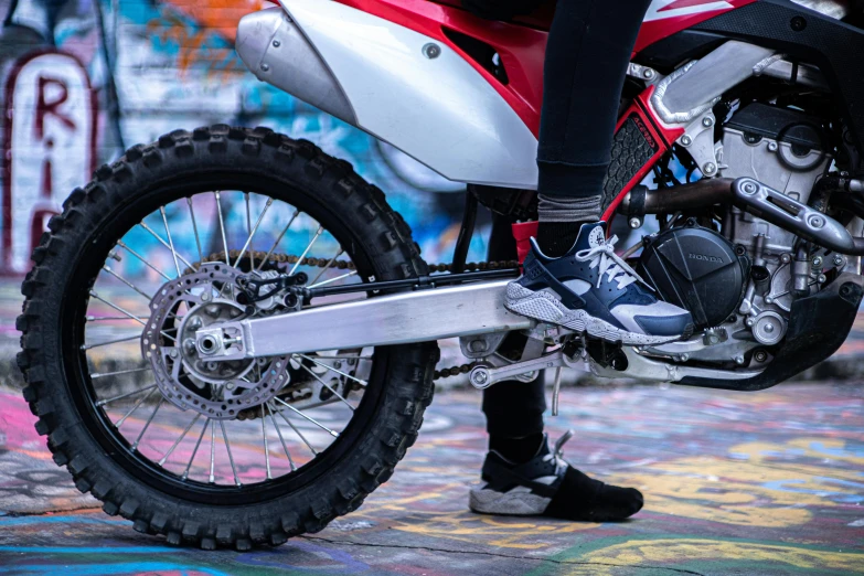 a person sitting on a motorcycle in front of a graffiti covered wall, a picture, unsplash, photorealism, red shell. dirt track, sneaker made out of lego, silver red white details, close-up on legs