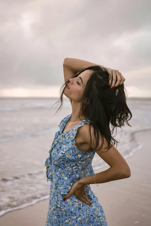 a woman standing on top of a beach next to the ocean, by Carey Morris, pexels contest winner, renaissance, long windy hair style, brunette, patterned clothing, blue