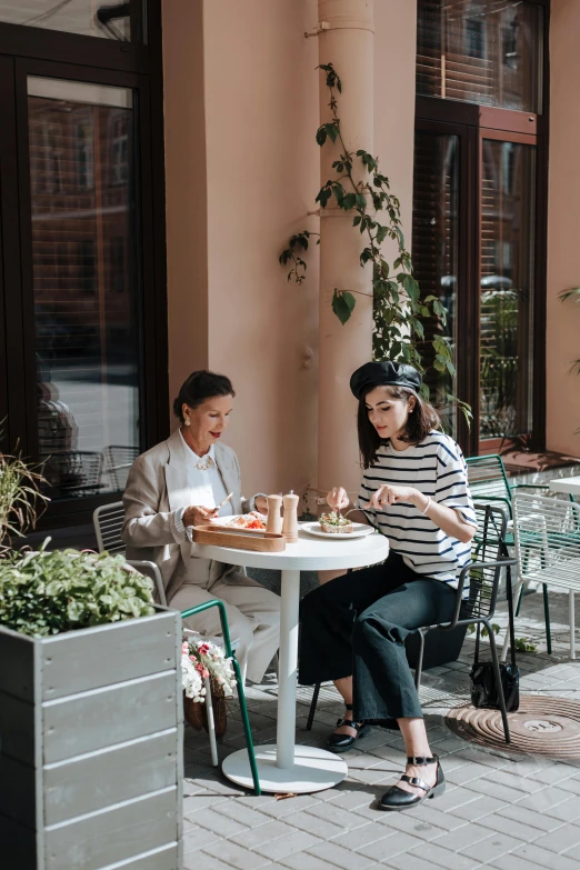 two women sitting at a table outside of a building, by Julia Pishtar, pexels contest winner, breakfast, wide full body, cafe interior, wearing a french beret