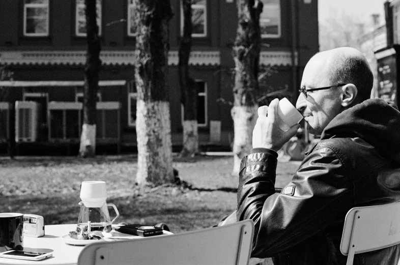 a black and white photo of a man sitting at a table, a black and white photo, by Aleksandr Gerasimov, pixabay, at the park, drinking coffee, bald man, gopnik in a black leather jacket