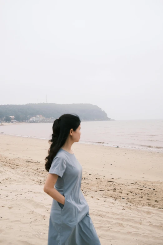 a woman standing on top of a sandy beach, inspired by Zhang Kechun, unsplash, song hye - kyo, wearing a t-shirt, blue gray, long distance