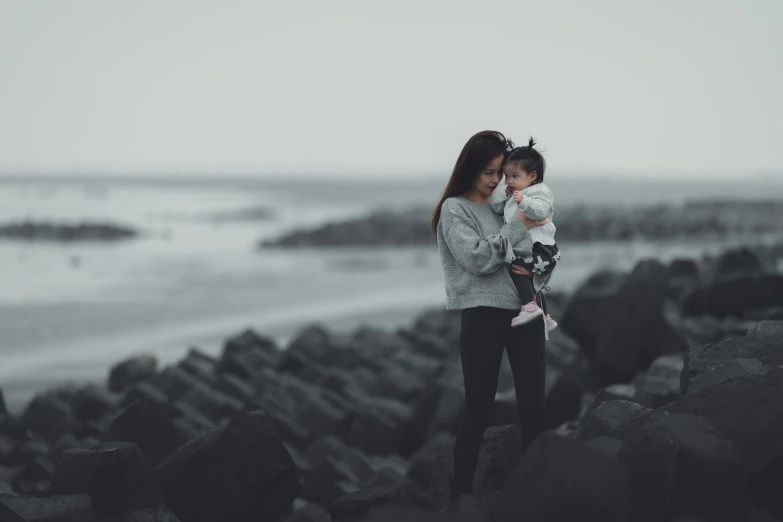 a woman holding a baby on top of a rocky beach, pexels contest winner, realism, grey and dark theme, looking cute, fashionable, slightly pixelated